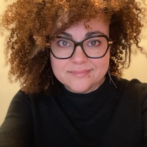 Woman with curly hair and glasses wearing a black turtleneck.